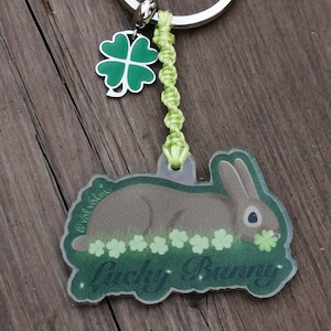 Peut inclure: Un porte-clés en forme de trèfle à quatre feuilles vert attaché à une chaîne verte avec un anneau de clé argenté. Le porte-clés présente un charme en plastique transparent avec un lapin brun couché sur un lit de trèfles verts et le texte "Lucky Bunny".