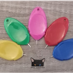Peut inclure: Cinq aiguilles à coudre en plastique de couleurs différentes avec un petit crochet métallique à l'extrémité. Les aiguilles sont disposées en demi-cercle sur une surface en bois. Les couleurs sont le vert, le rose, le rouge, le jaune et le bleu.