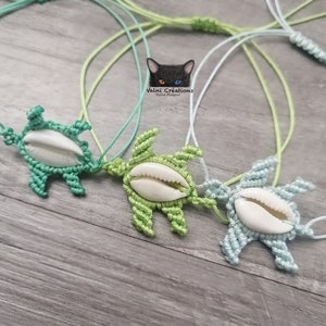 May include: Three macrame turtle necklaces in shades of green and blue, each with a white seashell centre. The necklaces are displayed on a wooden surface, with the Valni Créations logo visible in the background.