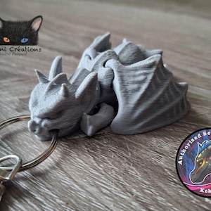 May include: A grey 3D-printed dragon keyring. The dragon has detailed wings, horns, and a fierce expression. The keyring includes a metal ring and clasp. The image also shows a logo with a cat and the text "Valni Créations".