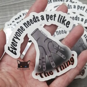 Peut inclure: Autocollant blanc avec du texte noir qui dit "Tout le monde a besoin d'un animal de compagnie comme 'La Chose'". Une main de dessin anim&eacute; grise cousue est repr&eacute;sent&eacute;e sous le texte. L'autocollant est tenu dans une main.