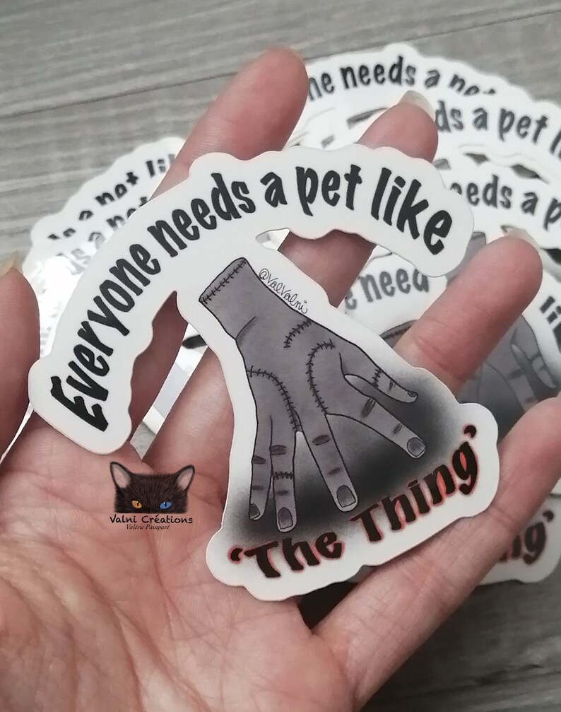 Peut inclure: Autocollant blanc avec du texte noir qui dit "Tout le monde a besoin d'un animal de compagnie comme 'La Chose'". Une main de dessin anim&eacute; grise cousue est repr&eacute;sent&eacute;e sous le texte. L'autocollant est tenu dans une main.