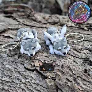 May include: Two gray and white wolf keychains with silver key rings are displayed on a textured, brown surface. The wolves are in a playful pose. A small black cat face with different colored eyes is also visible. A circular logo with a horse is in the background.