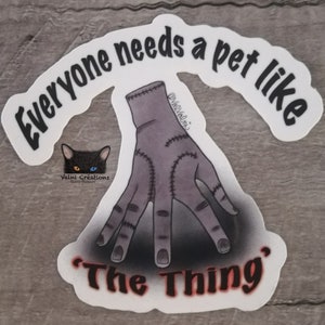 Peut inclure: Autocollant blanc avec du texte noir qui dit "Tout le monde a besoin d'un animal de compagnie comme 'La Chose'". Un chat noir avec des yeux bleus et orange est en arri&egrave;re-plan. Une main grise avec des points de suture est au premier plan.