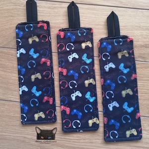 May include: Three black fabric bookmark with a gamer theme. The bookmarks feature a repeating pattern of colorful video game controllers and headphones. The bookmarks have black straps at the top.