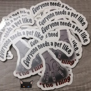 Peut inclure: Une pile d'autocollants blancs avec une illustration noire et grise d'une main avec le texte "Everyone needs a pet like The Thing" dessus.