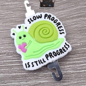 May include: A green snail with a pink heart and the text "Slow Progress is Still Progress" on a white background. The snail is attached to a silver hook.