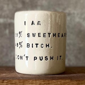 Può includere: Un bicchiere in ceramica color crema con testo nero che recita "I AM 81% SWEETHEART 19% BITCH. DON'T PUSH IT." Il bicchiere ha una forma cilindrica e una superficie leggermente strutturata.