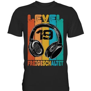 Può includere: Maglietta nera con una grafica retrò di cuffie con il numero 19 all'interno. Il testo "LEVEL" e "FREIGESCHALTET" è anche sulla maglietta.