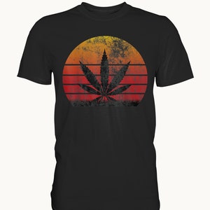 Könnte beinhalten: Ein schwarzes T-Shirt mit einem Vintage-Grafikdruck eines Marihuanablattes vor einem roten, orangefarbenen und gelben Sonnenuntergang.