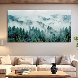 Könnte beinhalten: Ein großer rechteckiger Leinwanddruck, der eine neblige Waldszene zeigt. Das Kunstwerk zeigt einen dichten Wald aus grünen Bäumen mit einem Hintergrund aus Bergen, die in Nebel gehüllt sind. Ein Vogelschwarm fliegt über den Himmel. Der Druck wird über einem beigen Sofa angezeigt.