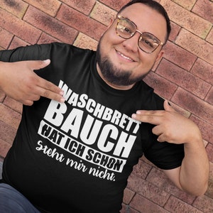May include: Black t-shirt with the German text "Waschbrett Bauch Hat Ich Schon Steht mir nicht" in white, worn by a person. The person is smiling and pointing at the text. The background is a brick wall.