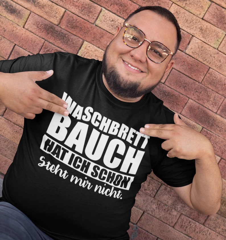 May include: Black t-shirt with the German text "Waschbrett Bauch Hat Ich Schon Steht mir nicht" in white, worn by a person. The person is smiling and pointing at the text. The background is a brick wall.