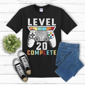 May include: Black t-shirt featuring a retro-style video game controller graphic and the text "LEVEL 20 COMPLETE".
