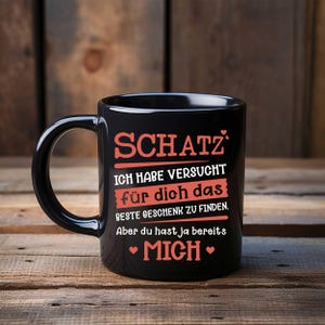 Könnte beinhalten: Schwarze Keramiktasse mit einem weißen und roten Design. Die Tasse trägt die Aufschrift "SCHATZ ICH HABE VERSUCHT für dich das BESTE GESCHENK ZU FINDEN. Aber du hast ja bereits • MICH •"