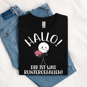May include: Black t-shirt featuring the word "HALLO!" in white, and a cartoon figure holding a pink brain. The text "DIR IST WAS RUNTERGEFALLEN!" is printed below the figure. The shirt is folded on top of blue denim jeans.