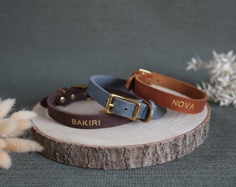 May include: Three leather dog collars with gold buckles and personalized names. The collars are brown, gray, and tan. The names on the collars are "Bakiri", "Nova", and "Nova".