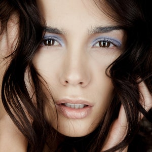 May include: A woman with long, dark brown hair, wearing light blue eyeshadow and nude lipstick. Her hair is covering part of her face.