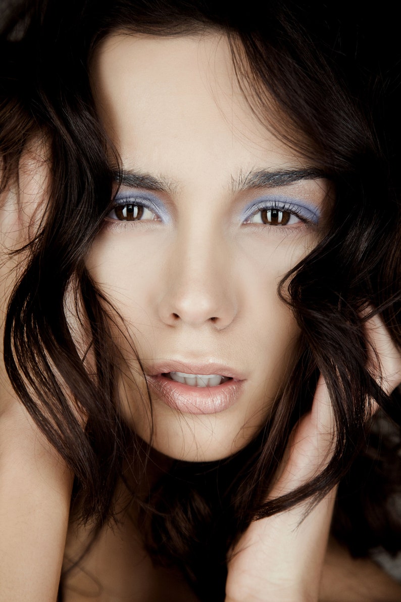 May include: A woman with long, dark brown hair, wearing light blue eyeshadow and nude lipstick. Her hair is covering part of her face.