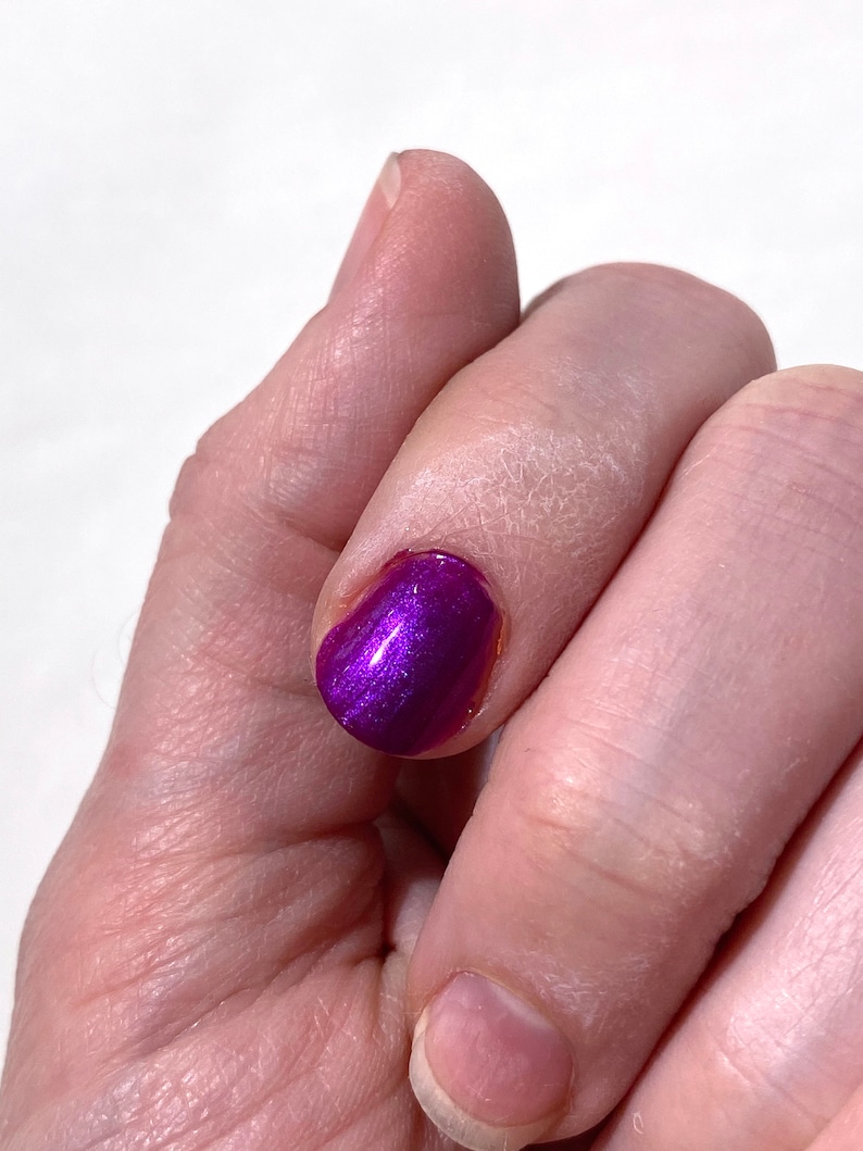 Puede incluir: Primer plano de un dedo con esmalte de u&ntilde;as morado con purpurina. El esmalte de u&ntilde;as es brillante y tiene un acabado met&aacute;lico.