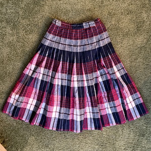 Puede incluir: Una falda plisada a cuadros en tonos de rojo, azul marino, blanco y gris. La falda presenta un patrón de cuadros clásico con pliegues verticales, creando una silueta completa en forma de A. La tela parece ser un material de peso medio.