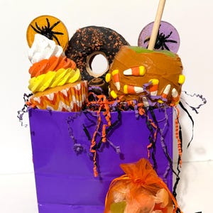 Puede incluir: Una bolsa de regalo morada llena de golosinas de Halloween. La bolsa contiene una magdalena, una manzana con caramelo decorada con caramelos de maíz, una rosquilla de chocolate y papel decorativo. Una pequeña bolsa naranja de caramelos está al frente.