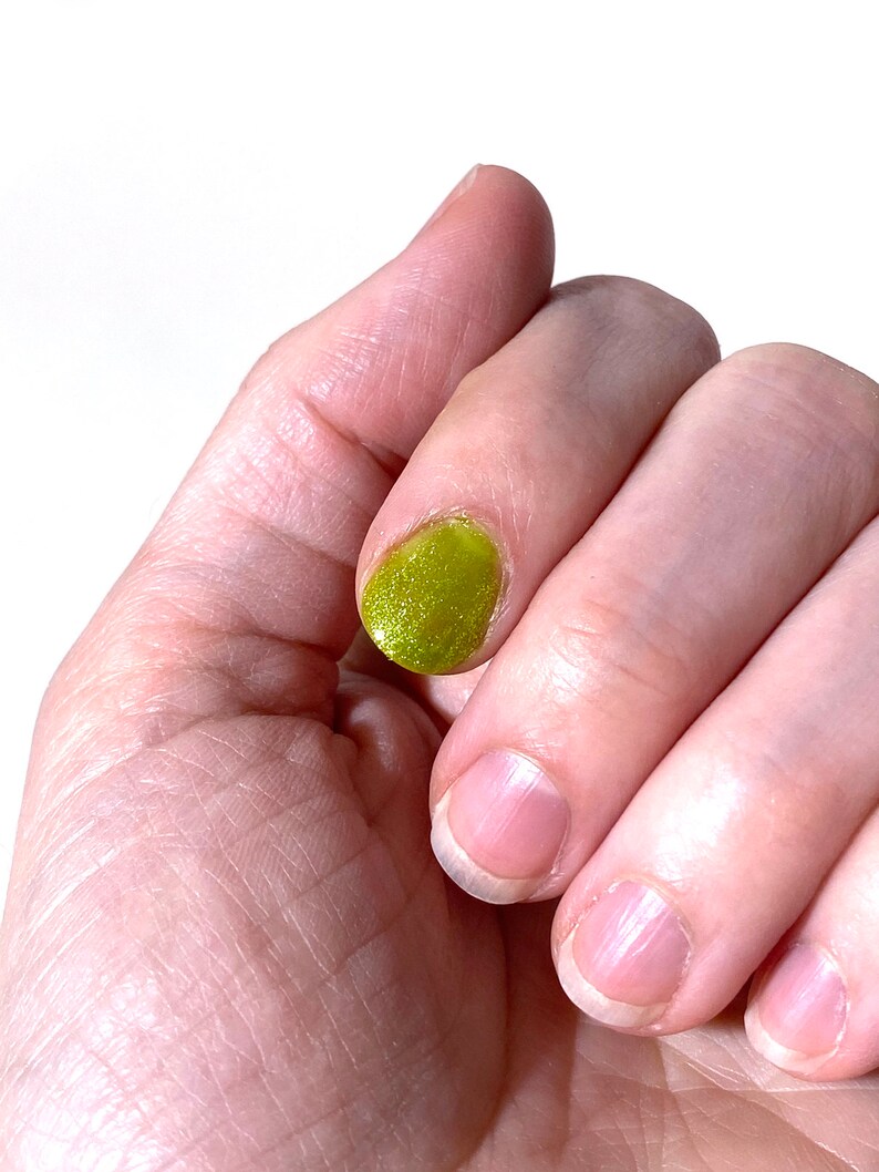 Puede incluir: Una mano con cuatro u&ntilde;as, una de las cuales est&aacute; pintada con un esmalte de u&ntilde;as verde brillante.
