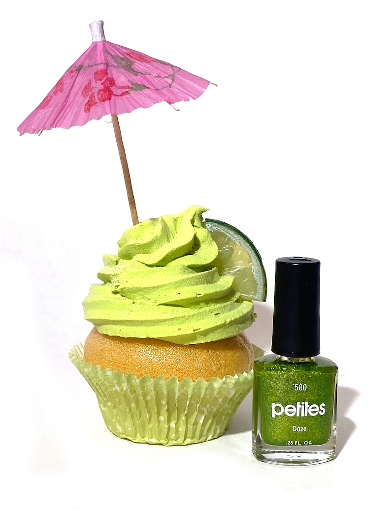 Puede incluir: Un cupcake verde con glaseado de lima y una rodaja de lima, coronado con una sombrilla de papel rosa. Una botella de esmalte de u&ntilde;as verde con purpurina est&aacute; al lado del cupcake. La etiqueta de la botella dice "pelites 580 Daze 0.25 fl oz."
