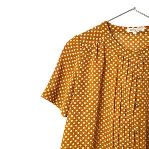 May include: Mustard yellow short-sleeved blouse with white polka dots. The blouse features a pleated front and small, round buttons. The garment is hanging on a silver hanger against a white background. The brand name "MISSEL" is visible on the label.