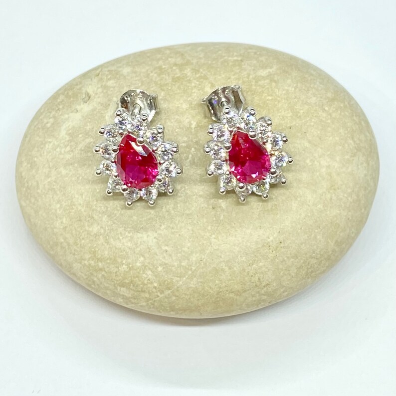 K&ouml;nnte beinhalten: Ein Paar silberne Ohrringe mit einem birnenf&ouml;rmigen roten Edelstein, der von kleineren klaren Edelsteinen umgeben ist.