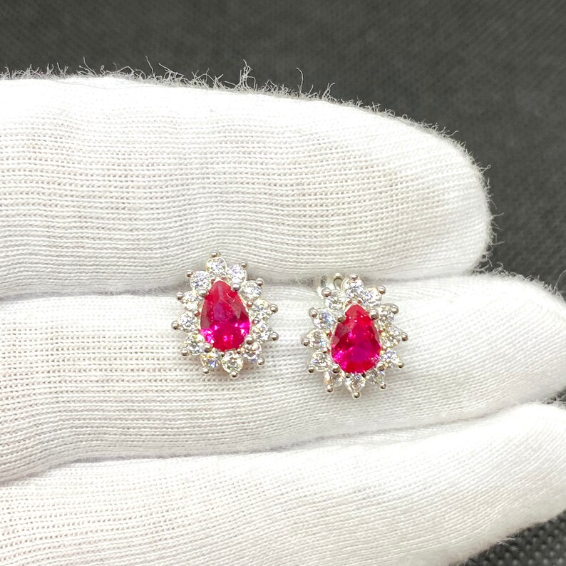 K&ouml;nnte beinhalten: Ein Paar silberne Ohrringe mit einem tropfenf&ouml;rmigen roten Edelstein, umgeben von kleineren klaren Edelsteinen.