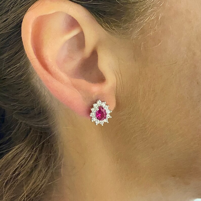 K&ouml;nnte beinhalten: Ein Paar silberne Ohrringe mit einem birnenf&ouml;rmigen roten Edelstein, der von einem Heiligenschein aus kleineren klaren Edelsteinen umgeben ist.