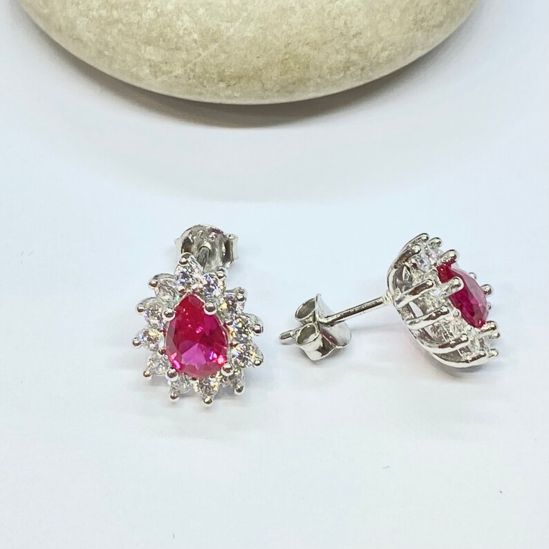 K&ouml;nnte beinhalten: Ein Paar silberne Ohrringe mit einem tropfenf&ouml;rmigen roten Edelstein, der von kleineren klaren Edelsteinen umgeben ist.