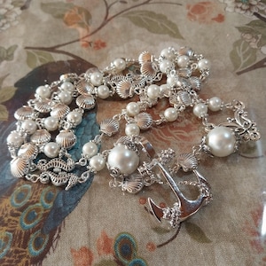 May include: A nautical-themed necklace featuring faux pearls, silver seashell charms, and an anchor pendant. The necklace has a delicate design with multiple strands of pearls and seashell accents. The anchor pendant adds a maritime touch to the jewelry.