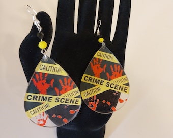 May include: A pair of dangle earrings with a black and yellow crime scene tape design. The earrings are shaped like teardrops and have red handprints on them. The text on the earrings reads "CAUTION! CRIME SCENE CAUTION!"