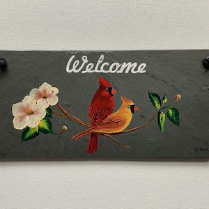 May include: A rectangular slate welcome sign featuring a hand-painted design. The sign displays the word "Welcome" in white script above a branch with two cardinals and white flowers. The birds are red and yellow.