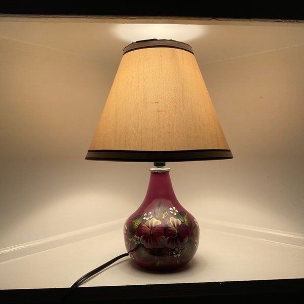 Hand painted colorful pansies on dark rose glass bottom lamp with a lampshade on top.