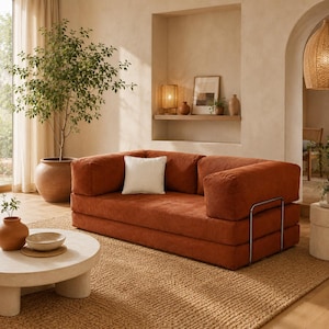 Op de afbeelding: Een roestkleurige corduroy bank met een wit vierkant kussen. De bank heeft een laag profiel en een metalen frame. Hij staat op een geweven jute vloerkleed. Een grote plant in pot en een ronde salontafel staan ook in de kamer.