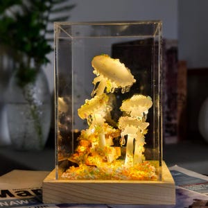 Könnte beinhalten: Ein klarer Glaswürfel mit einer Holzbasis, der eine Harzskulptur von drei leuchtenden Pilzen mit einem weißen und gelben Farbschema enthält. Die Pilze sind von einem Bett aus buntem Glitzer umgeben.