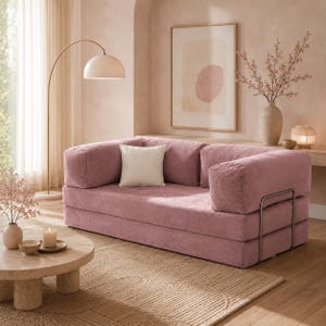 Op de afbeelding: Een stoffen roze corduroy bank met een wit vierkant kussen. De bank heeft een laag profiel en een metalen armleuning. Een ronde salontafel, een vloerlamp en een ingelijst abstract kunstwerk staan ook in de kamer.