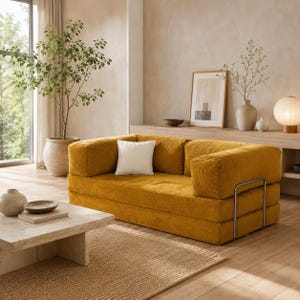 Op de afbeelding: Een mosterdgele corduroy bank met een wit vierkant kussen. De bank heeft een modern design met een metalen frame en staat in een woonkamer. Een neutraal tapijt en een marmeren salontafel staan voor de bank.