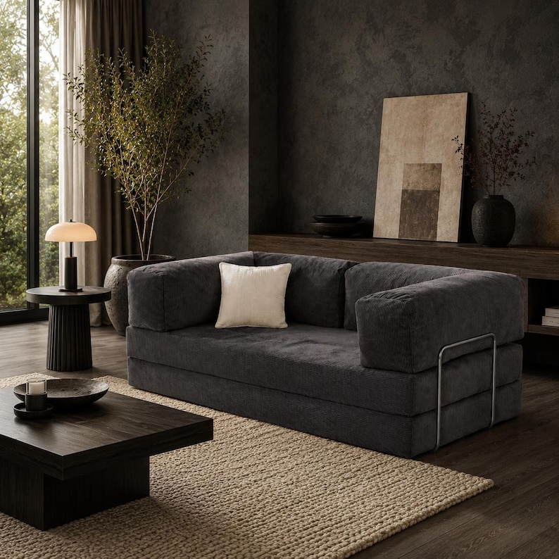 Op de afbeelding: Een donkergrijze modulaire bank met een wit vierkant kussen, in een moderne woonkamer. De bank heeft een ribfluweel textuur en metalen armleuningen. Een beige tapijt en donkere houten meubels maken de sc&egrave;ne compleet.
