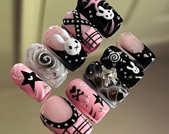 May include: A set of ten artificial nails with various designs. The nails feature a mix of pink, black, and clear colors, with embellishments like stars, skulls, and bunny designs. Some nails have a French tip design with black accents.