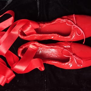 May include: A pair of red satin ballet slippers with matching red satin ribbon ties. The slippers are tied together with the ribbon.