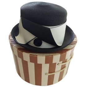 Puede incluir: Sombrero de paja negro con ala ancha y corona cilíndrica, adornado con una cinta negra y blanca. El sombrero descansa sobre una caja de sombreros vintage a rayas con asas de cuerda. La caja es marrón y crema.