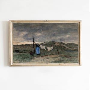 Op de afbeelding: Een antiek ingelijst aquarel schilderij toont een vrouw die wasgoed ophangt aan een waslijn in een veld. Het kunstwerk heeft een gedempt kleurenpalet van blauw, groen en grijs, met een bewolkte lucht en een verre heuvel.