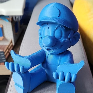 Peut inclure: Figurine bleue imprimée en 3D représentant un personnage de dessin animé assis, portant un chapeau et une salopette. La figurine est dans une pose détendue, les bras tendus et les pouces levés. La figurine est posée sur une surface grise.
