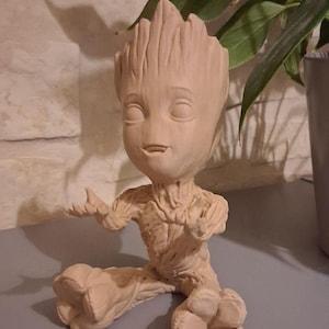 Peut inclure: Une figurine en céramique beige représentant Groot, un personnage du film Les Gardiens de la Galaxie, assis, les bras tendus et les jambes croisées. La figurine est détaillée avec une surface texturée et une expression amicale.