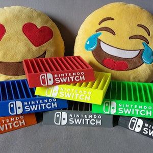 Peut inclure: Six supports de cartes de jeu Nintendo Switch en plastique de couleurs différentes. Chaque support a un logo Nintendo Switch blanc et les mots "Nintendo Switch" imprimés sur le devant. Les supports sont disposés en rangée sur une surface grise.