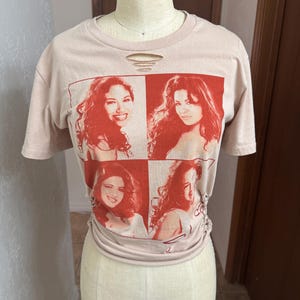 May include: A light pink t-shirt featuring a four-panel image of a woman with red hair. The image is printed in red on the front of the shirt. The shirt has a distressed look with a few small holes near the neckline.